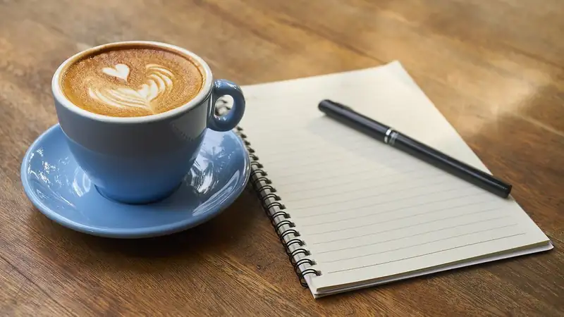 A latte with heart foam art in a blue cup beside a spiral notebook and pen on a wooden table.
