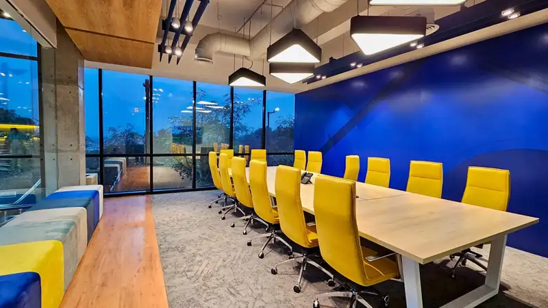 A modern conference room with a long wooden table, bright yellow chairs and a wall of windows at dusk.