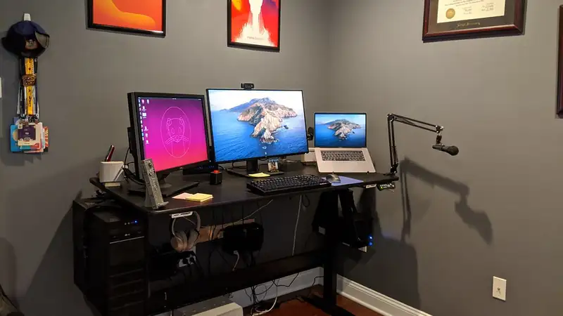 A home-office corner with a black desk, two monitors, a laptop and a microphone arm against a grey wall.