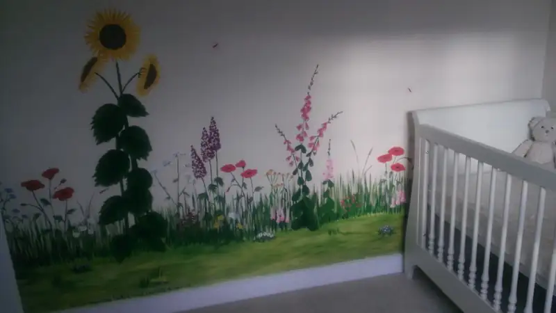 A white crib holding a teddy bear beside a nursery wall painted with sunflowers, poppies and wildflowers.