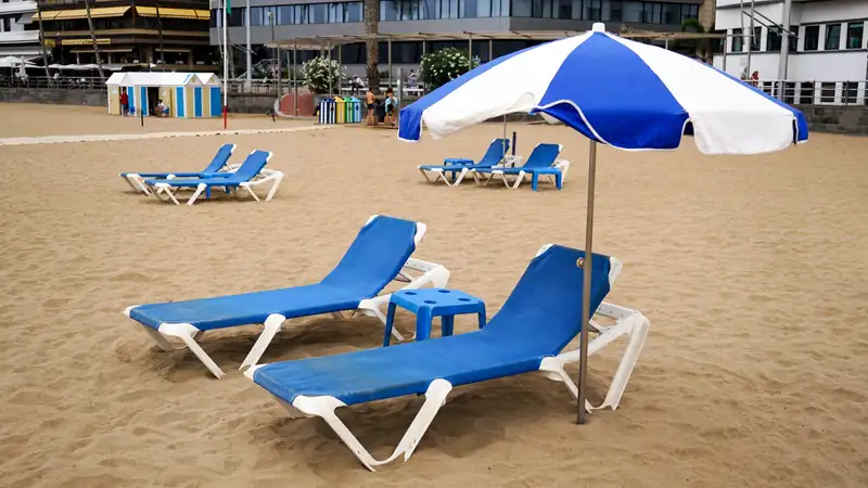 Two blue lounge chairs and a blue-and-white umbrella on a sandy city beach.