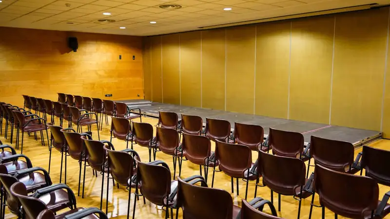 An empty wood-paneled conference room with rows of dark chairs facing a low stage.