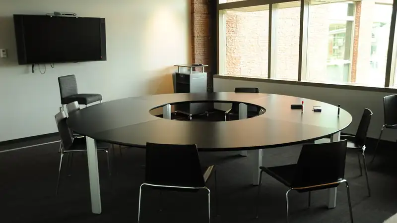 A round black conference table ringed by chairs in a bright meeting room with a wall-mounted screen.