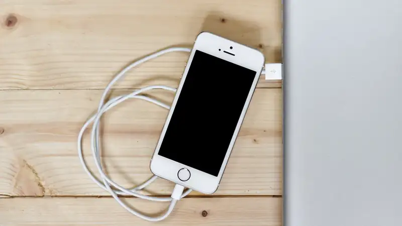 A white smartphone with its charging cable coiled beside it on a light wooden desk.