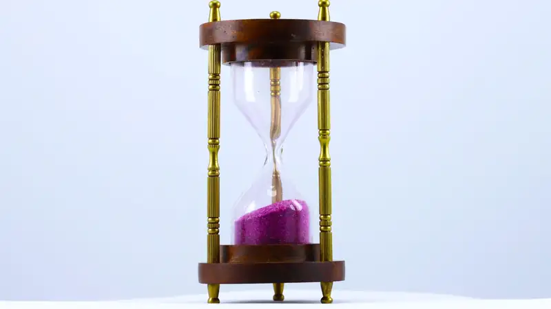 A brass-and-wood hourglass with magenta sand running through it against a pale blue backdrop.